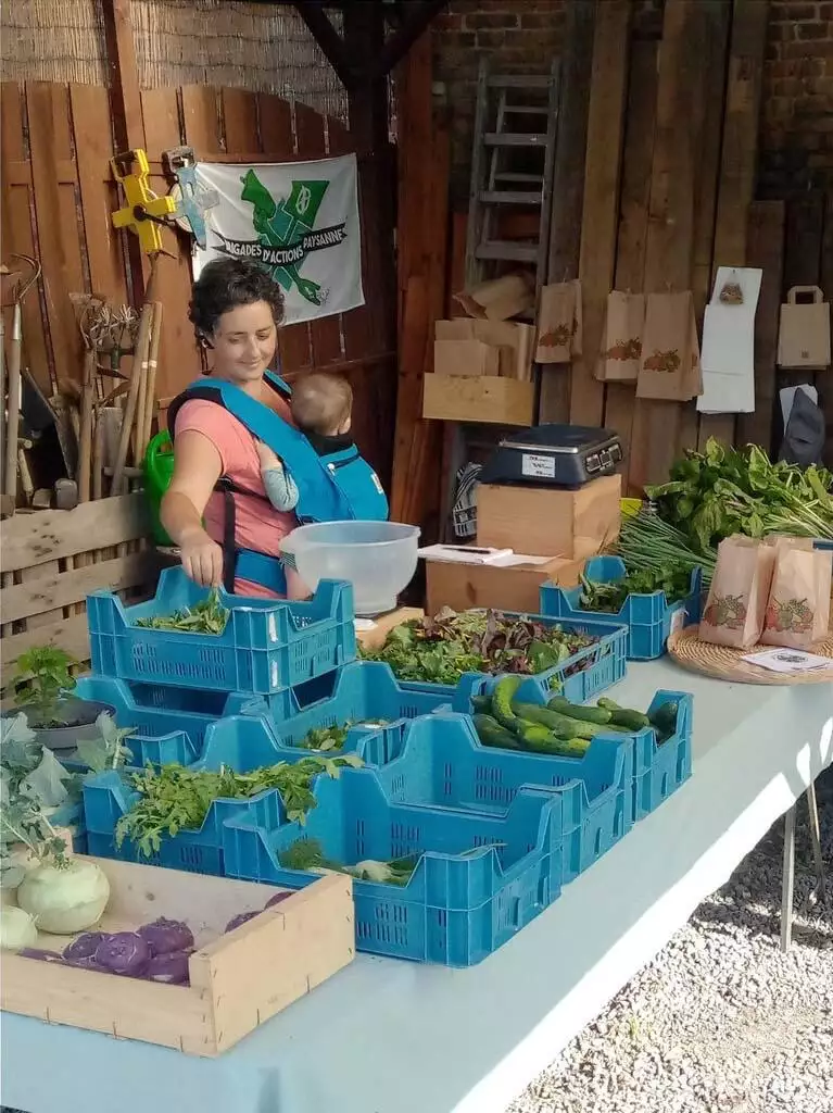 Une femme avec un bébé dans un porte-bébé fait ses courses chez un maraîcher avec des légumes frais disposés sur une table.